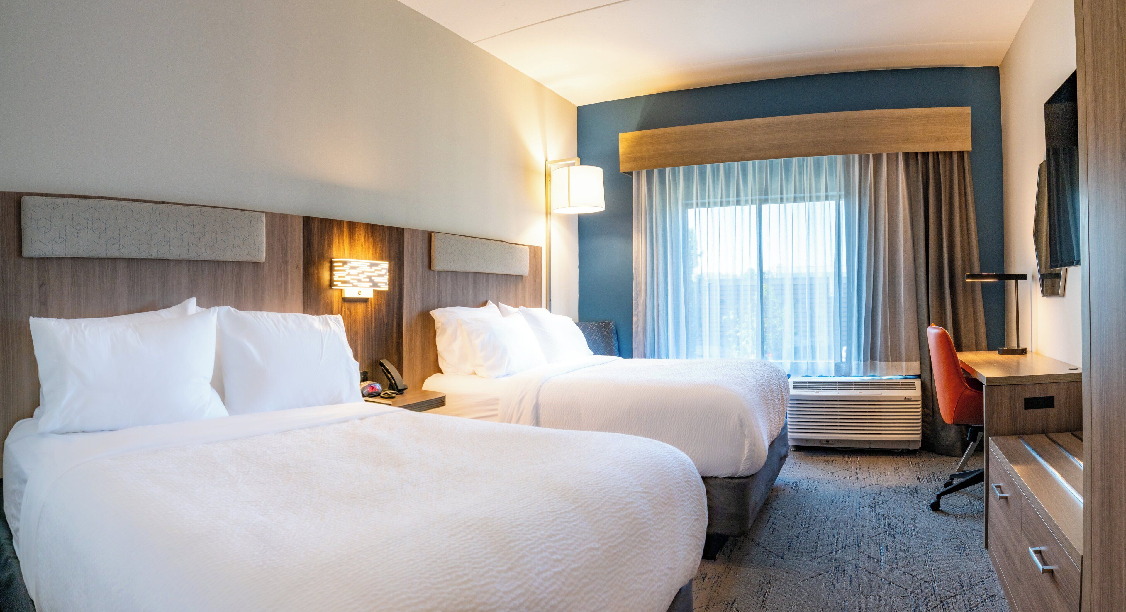 Une chambre d’hôtel avec deux lits, un bureau, une chaise, une télévision et une fenêtre avec des rideaux.