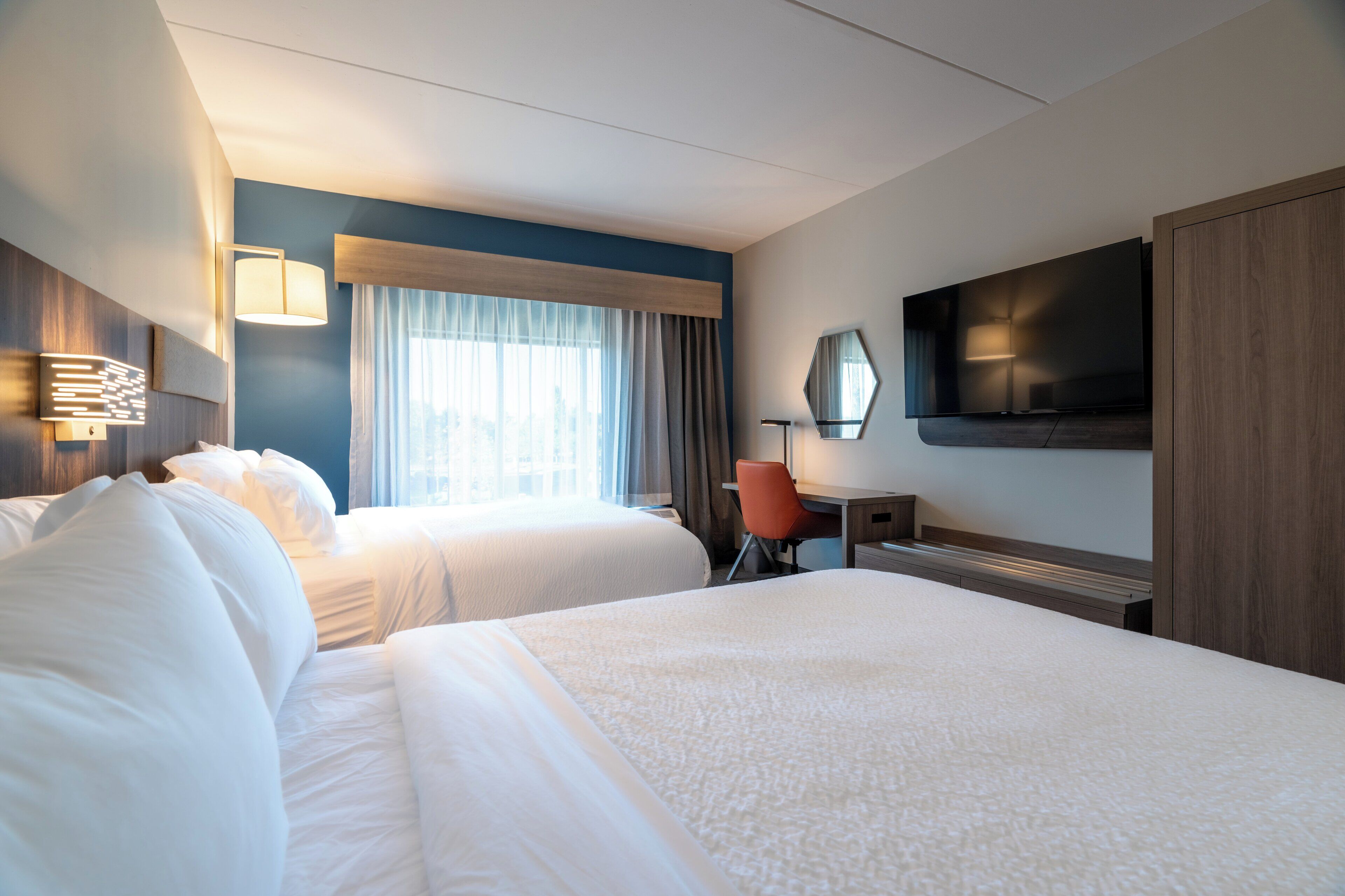 A hotel room with two beds, a desk, a chair, a TV, and a window with curtains.