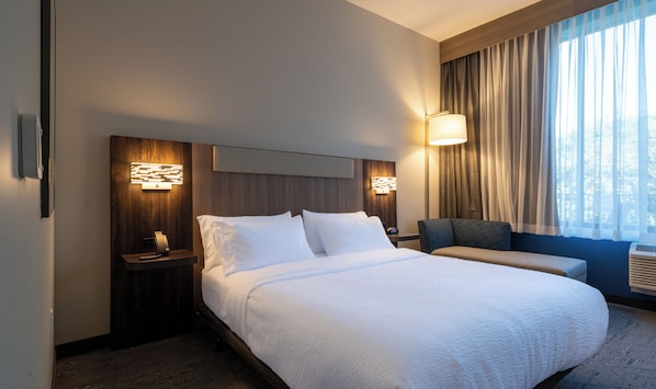 Ein Hotelzimmer mit einem großen Bett, einem Nachttisch mit Lampe, einem Sessel und einem Fenster mit Vorhängen.