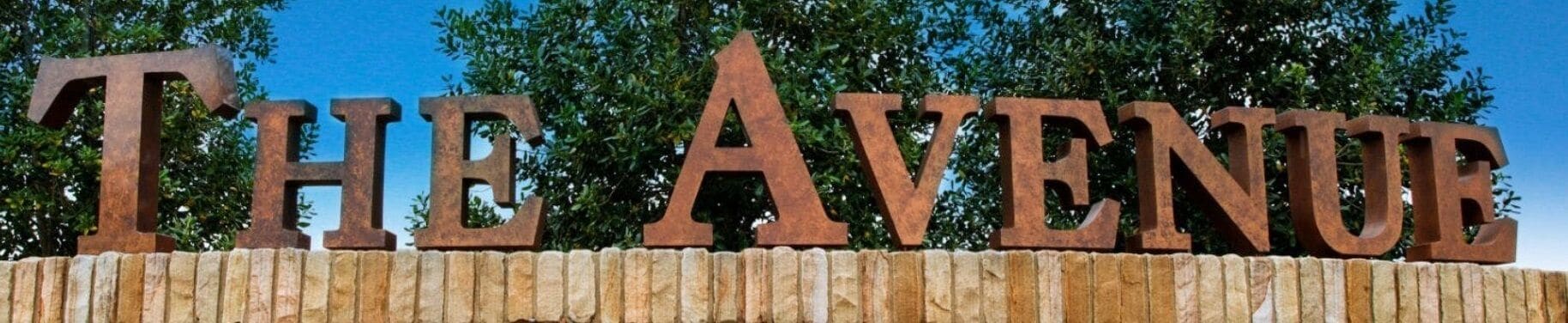 Een bord met de woorden 'THE AVENUE' in grote, verroeste metalen letters bevestigd aan een stenen muur.