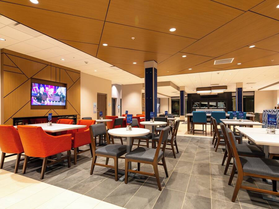 Eine moderne Hotelhalle mit mehreren Essplätzen, Tischen und Stühlen, einem großen Bildschirm und einer Rezeption.
