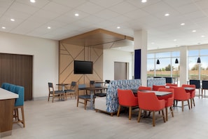 A modern indoor dining area with tables and chairs, a large screen, and a view through the windows. - Holiday Inn Express & Suites Prosper South - Frisco by IHG (Prosper)