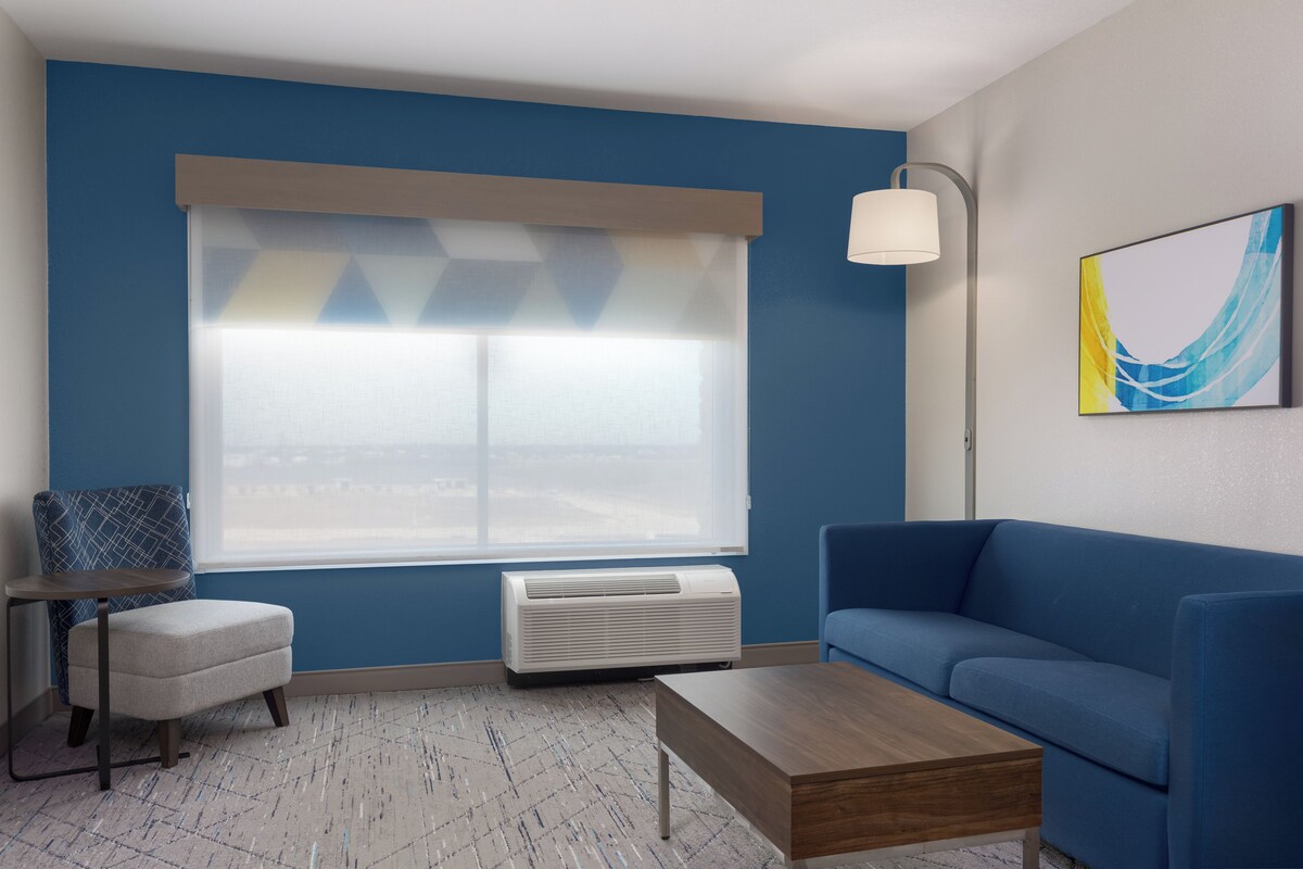 A room with a blue wall, a window with blinds, a blue sofa, a small table, a lamp, and a painting.