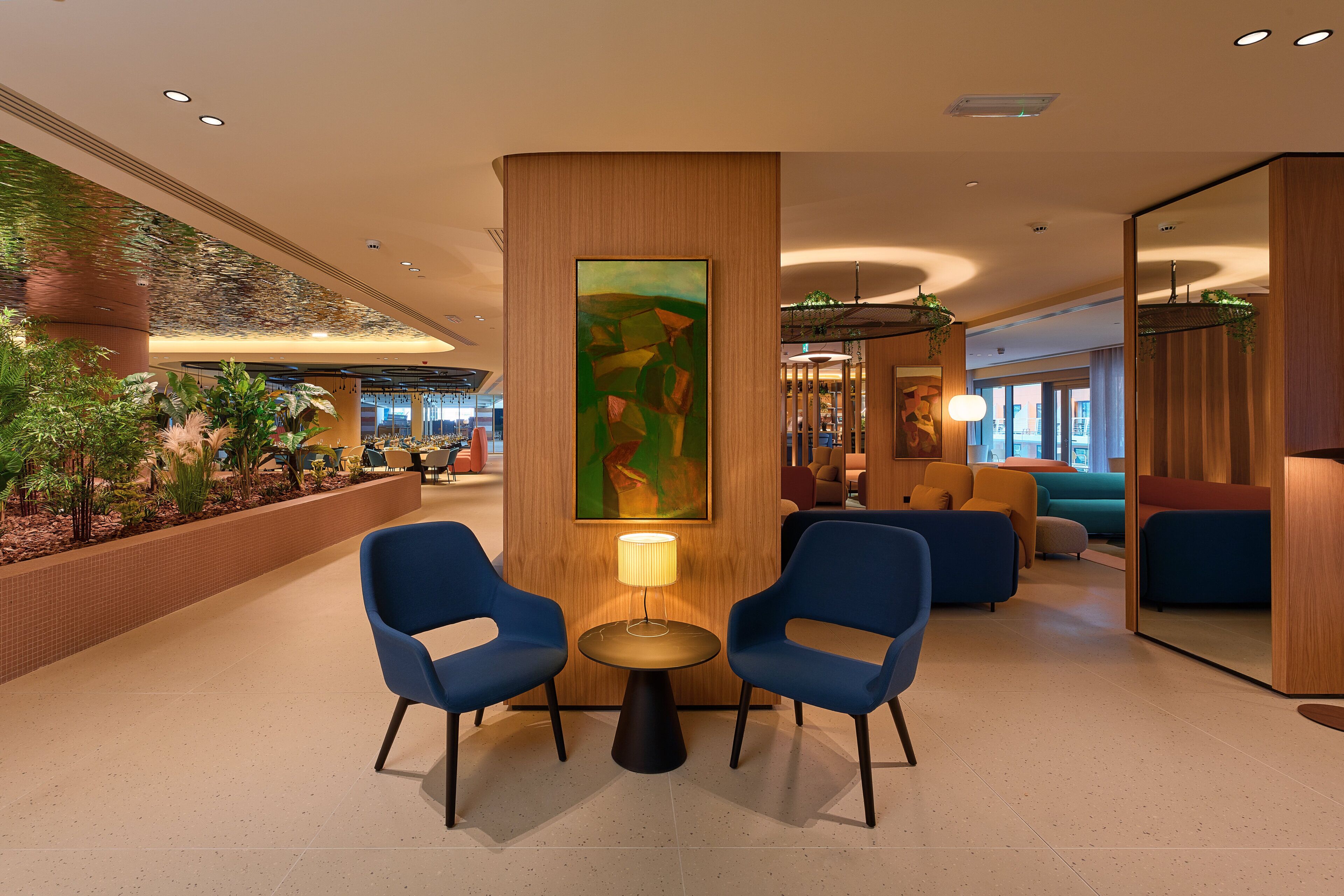 A lobby with two blue chairs around a small table with a lamp, a large abstract painting on a pillar, and a plant wall.