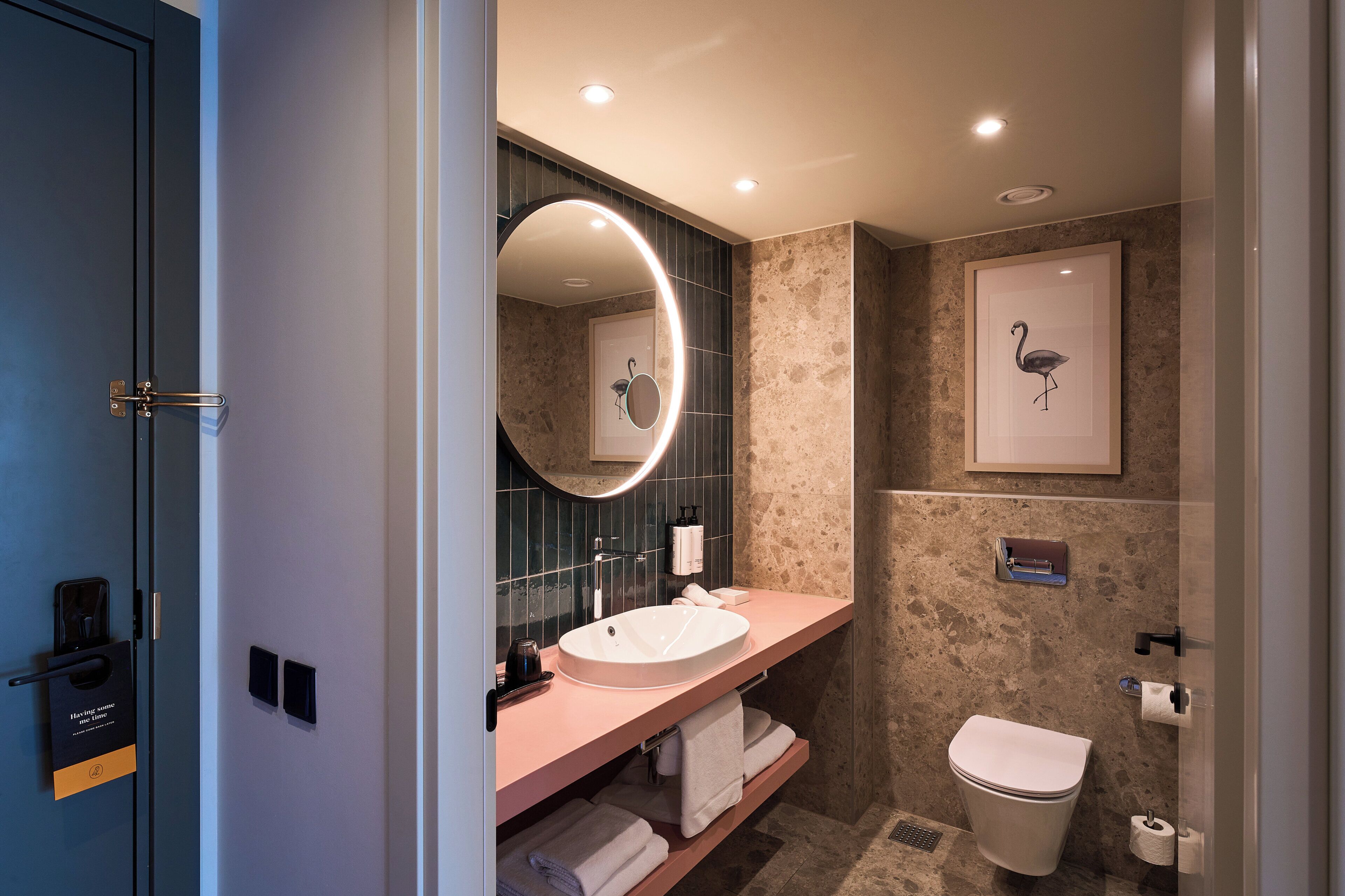 A modern bathroom with a round mirror, a sink, a toilet, and a framed artwork of a flamingo.