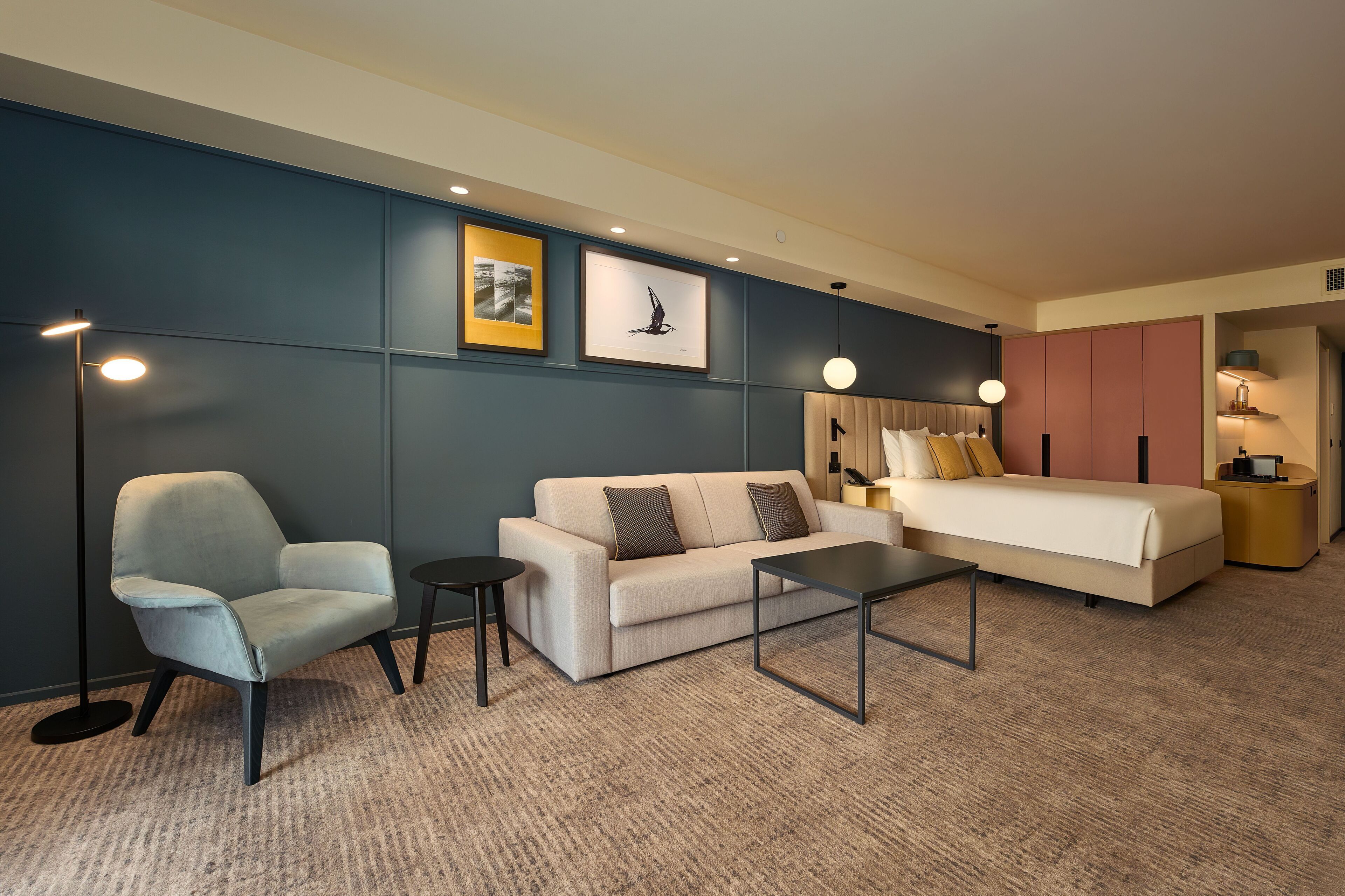 A hotel room with a sofa, armchair, coffee table, bed, bedside tables, and wall-mounted artwork.