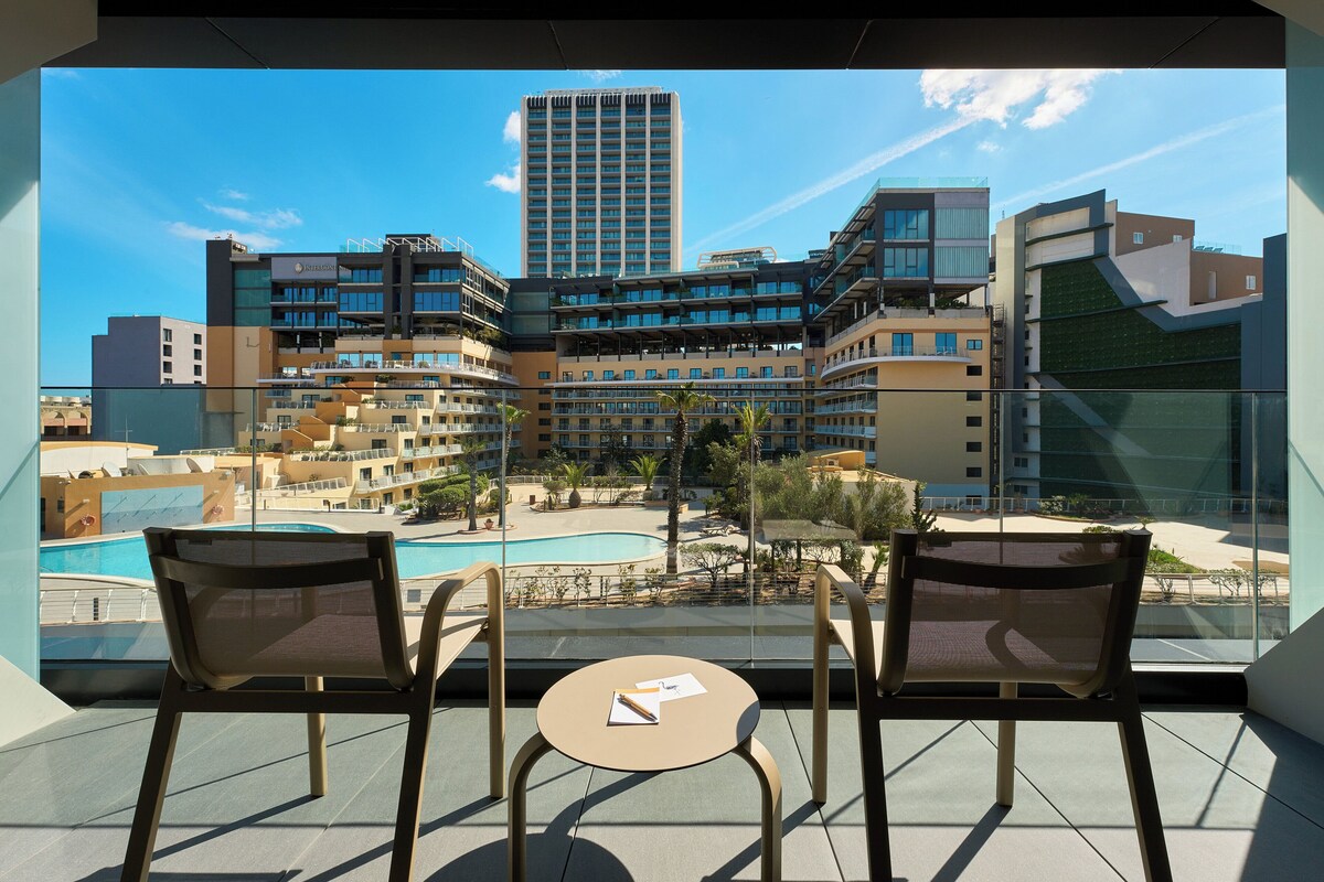 A balcony with two chairs and a small table, overlooking a swimming pool and a cityscape with modern buildings.