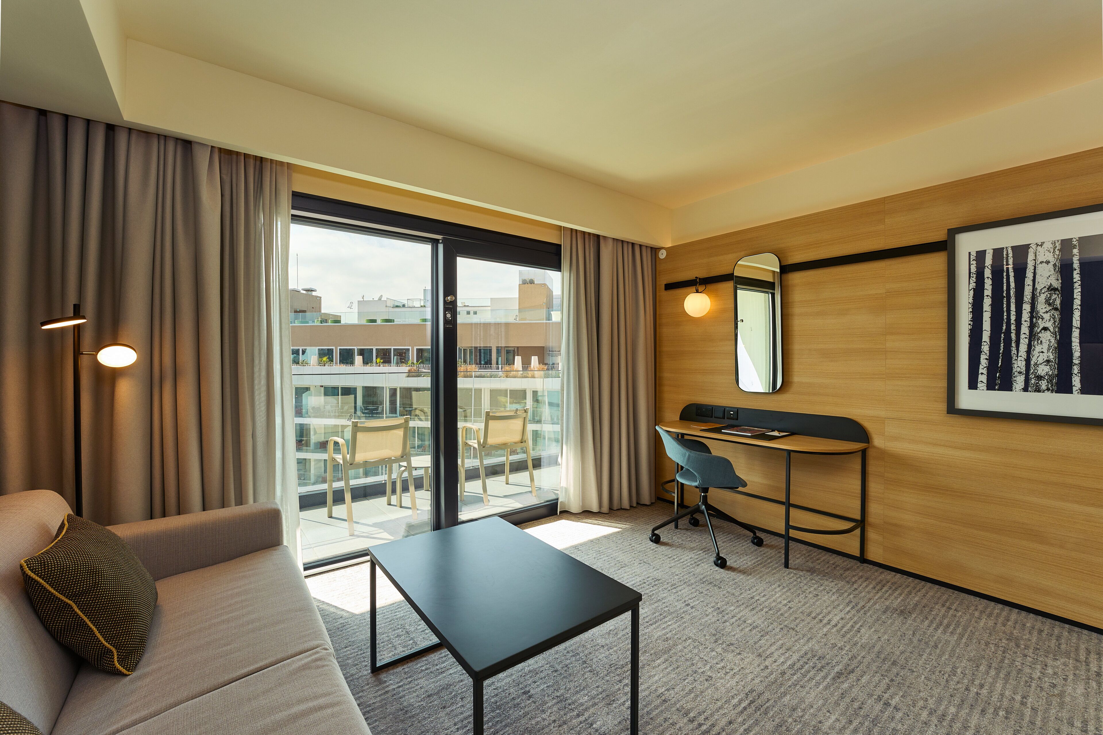 Habitación de hotel con sofá, escritorio, silla, mesita pequeña, ventana grande con cortinas, espejo en la pared y un cuadro enmarcado.