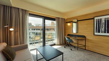 Habitación de hotel con sofá, escritorio, silla, mesita pequeña, ventana grande con cortinas, espejo en la pared y un cuadro enmarcado.
