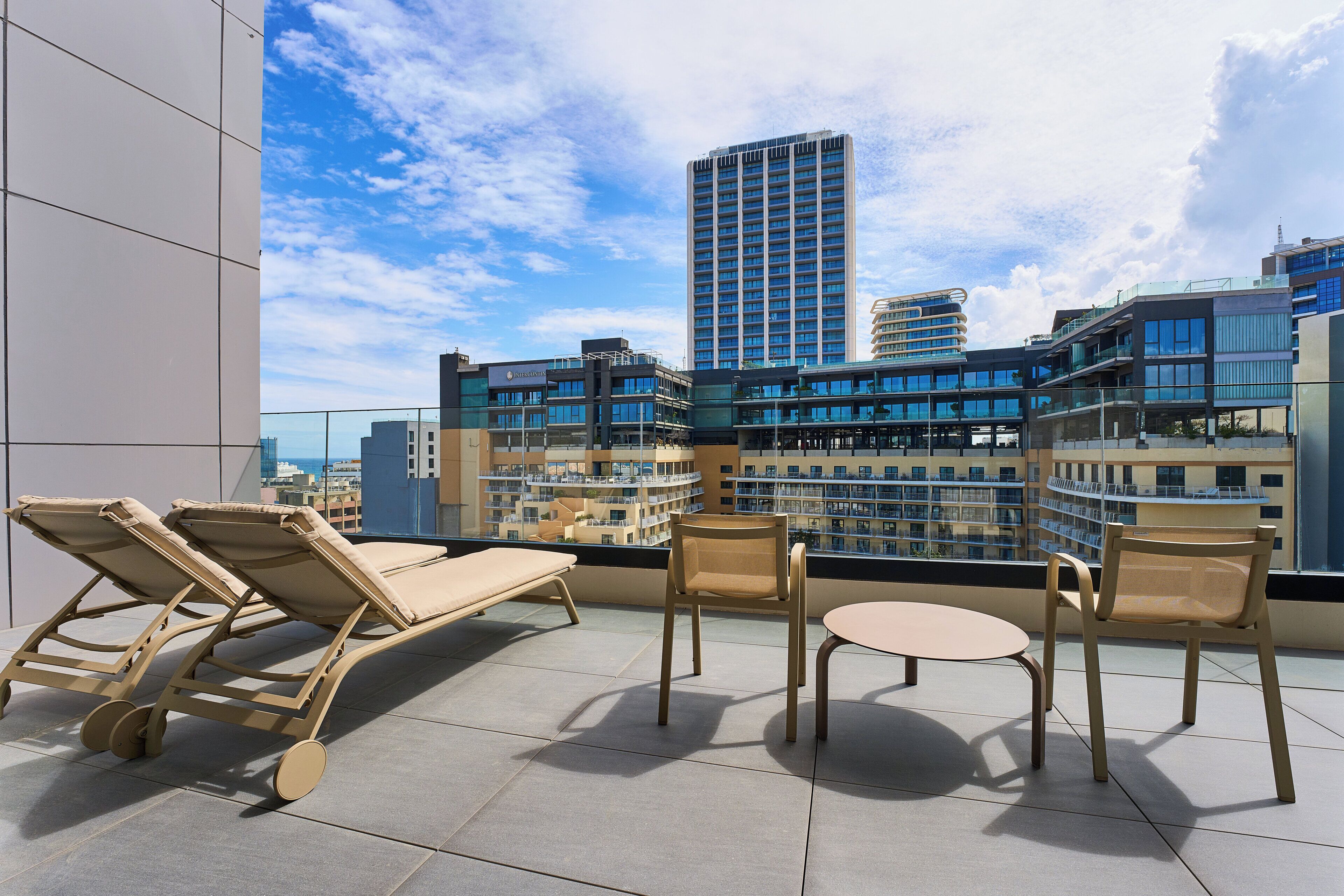A rooftop terrace with lounge chairs, a table, and a view of a cityscape with modern buildings.