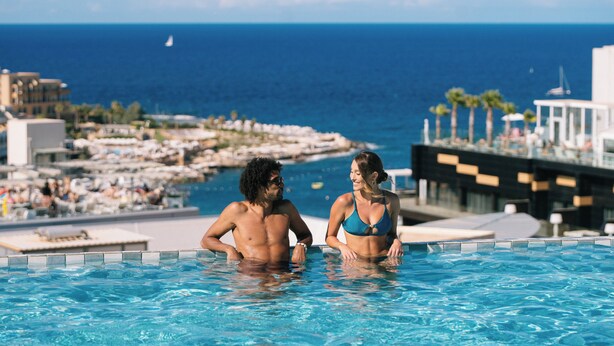 A couple in a swimming pool with a view of the ocean and resort buildings. - voco Malta by IHG (St. Julian's)