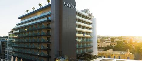 Un hotel moderno con un llamativo letrero 'Voco', varios balcones y una terraza en la azotea con tumbonas.