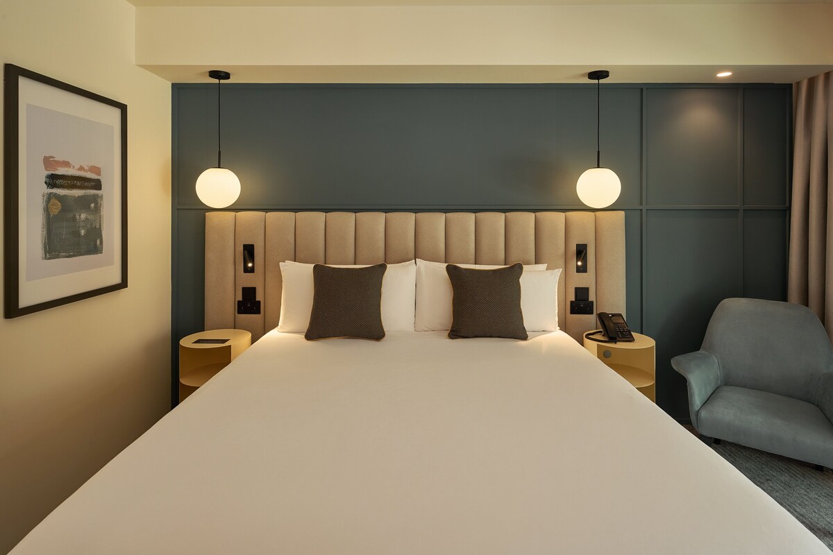 A modern hotel room with a large bed, two bedside tables, a chair, and a framed picture on the wall.