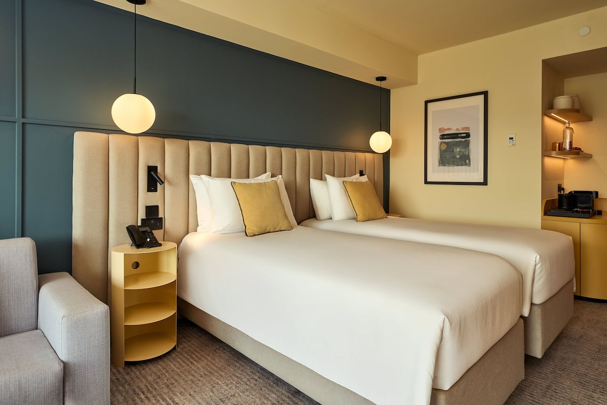 A hotel room with two beds, a desk, a chair, and a lamp.