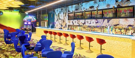 Zona de bar con sillas azules, una barra con botellas y un mural floral.