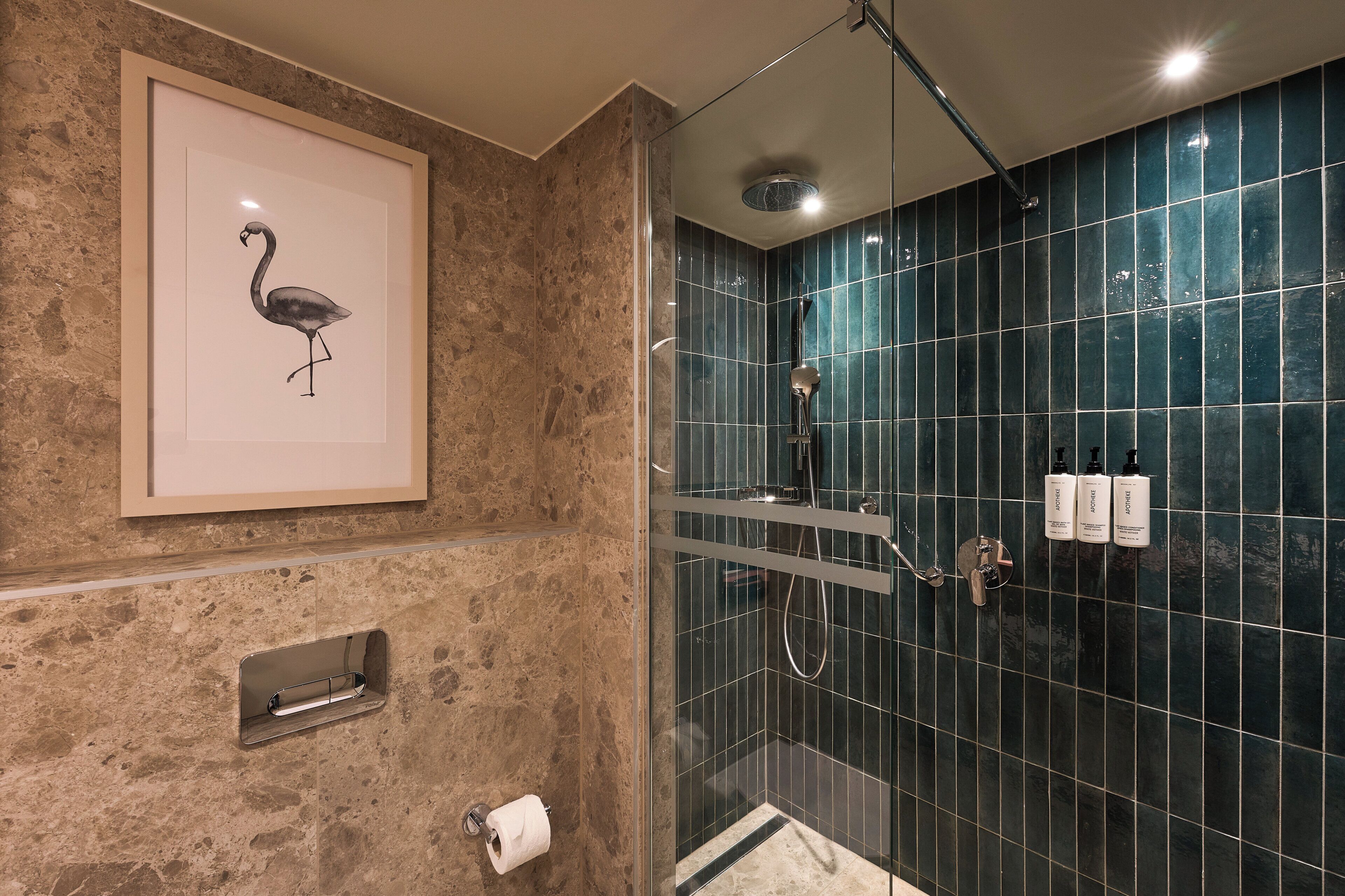 A bathroom with a framed artwork of a flamingo, a glass-enclosed shower with a rain showerhead, and a towel rack.