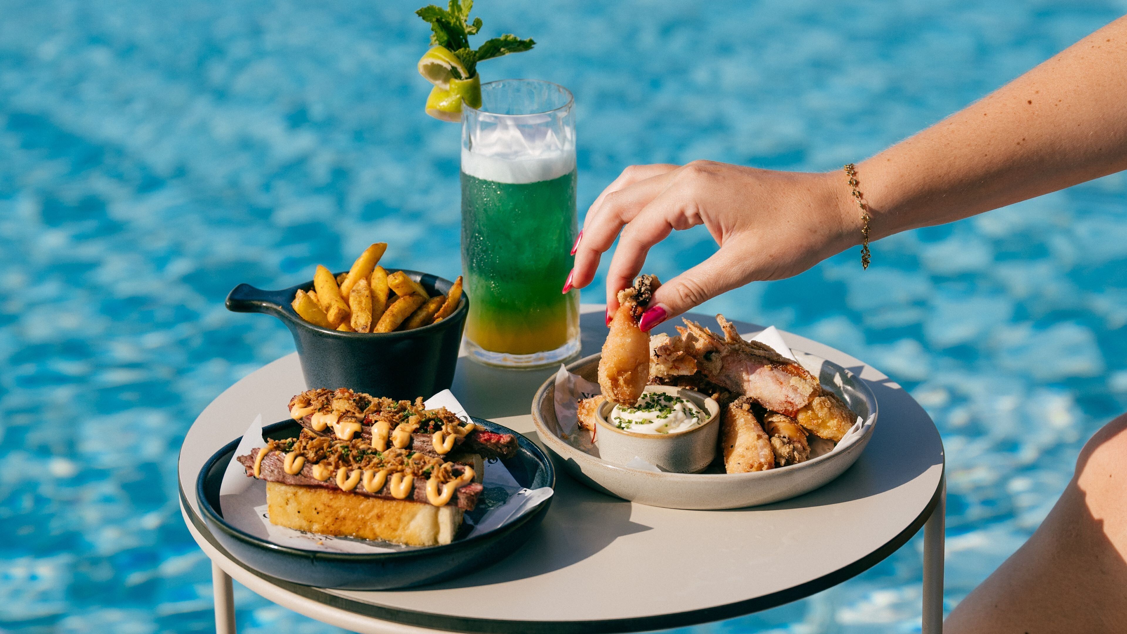 Una mesa con comida y bebidas junto a una piscina.