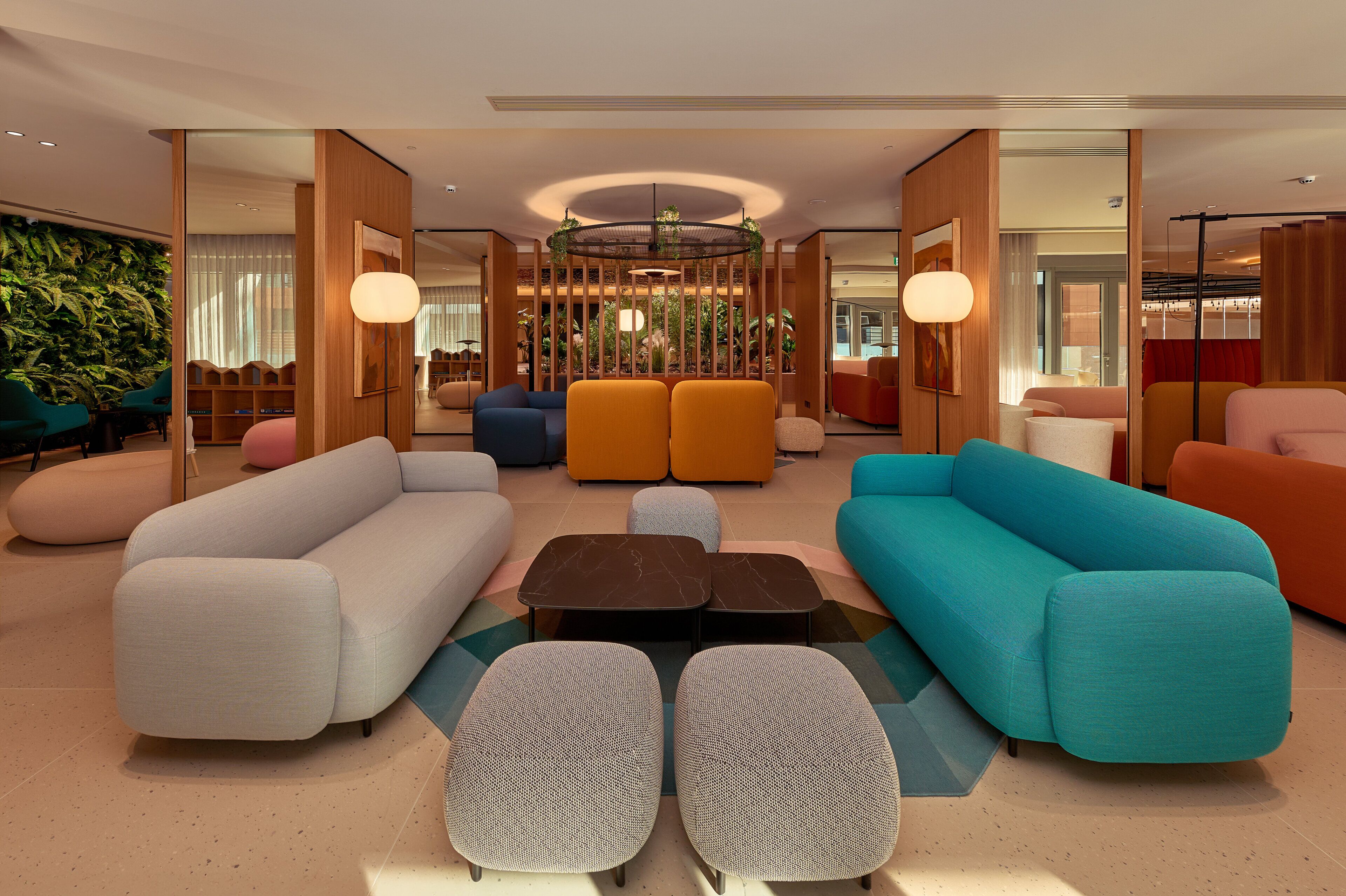 A modern hotel lobby with a central seating area featuring sofas and ottomans, a decorative wooden partition, and a large plant wall.
