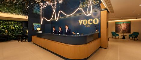 Una recepción con tres empleados, una pared con el logotipo de 'VOCO Malta' y una pared verde con plantas de fondo.