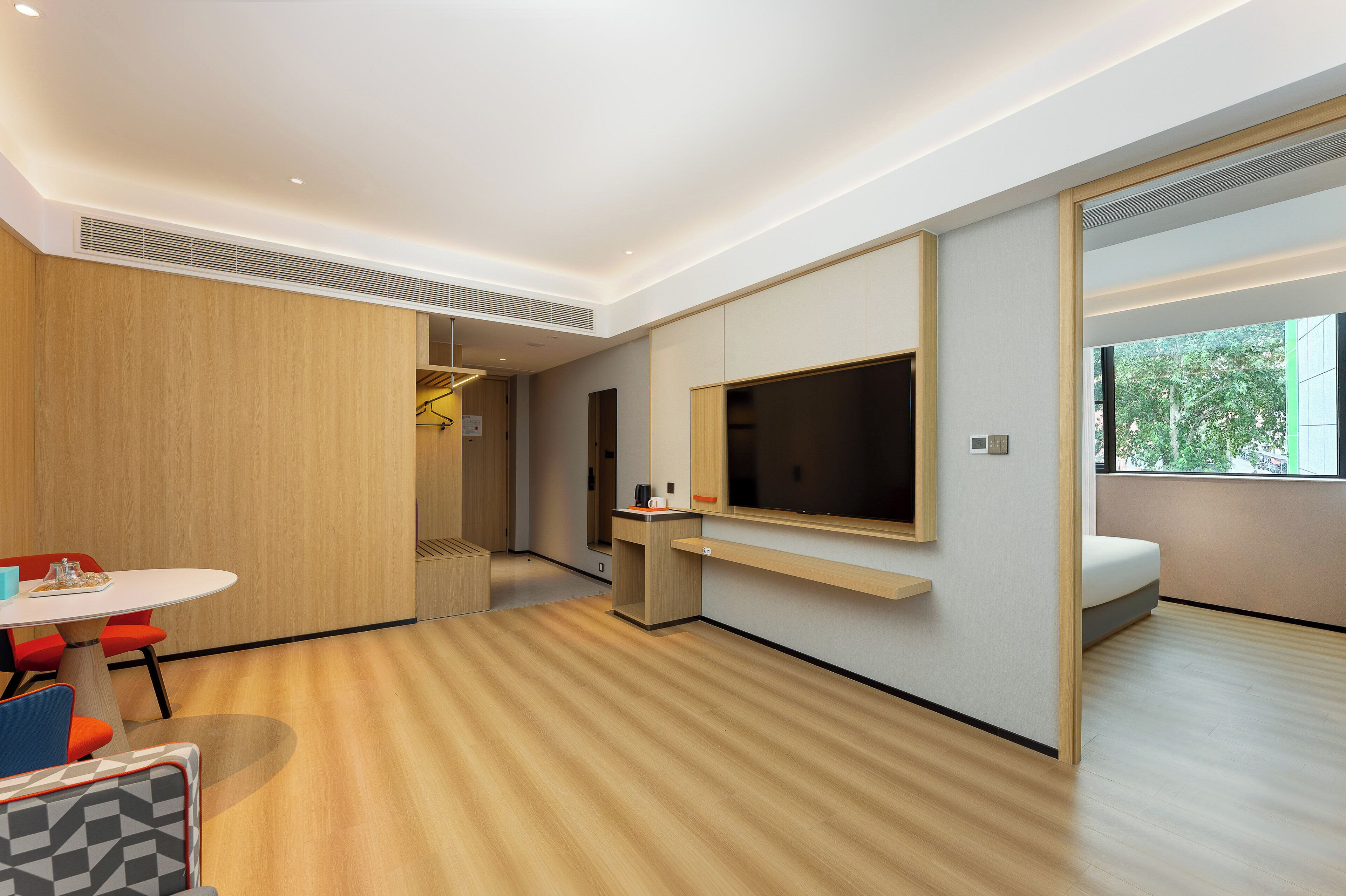 A modern hotel room with a wooden floor, a flat-screen TV mounted on the wall, a small desk with a chair, and a window offering a view of greenery.