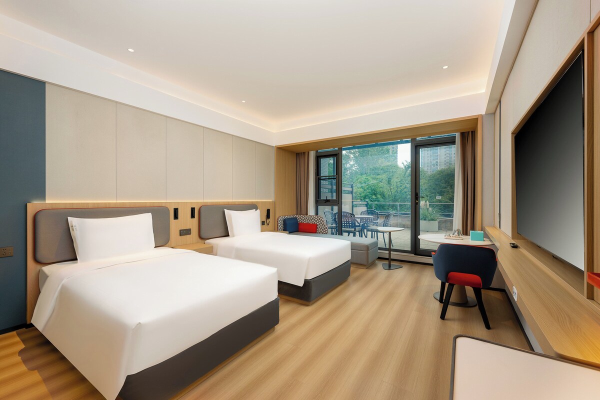 A hotel room with two beds, a small table, a chair, and a large window offering a view of the outdoors.