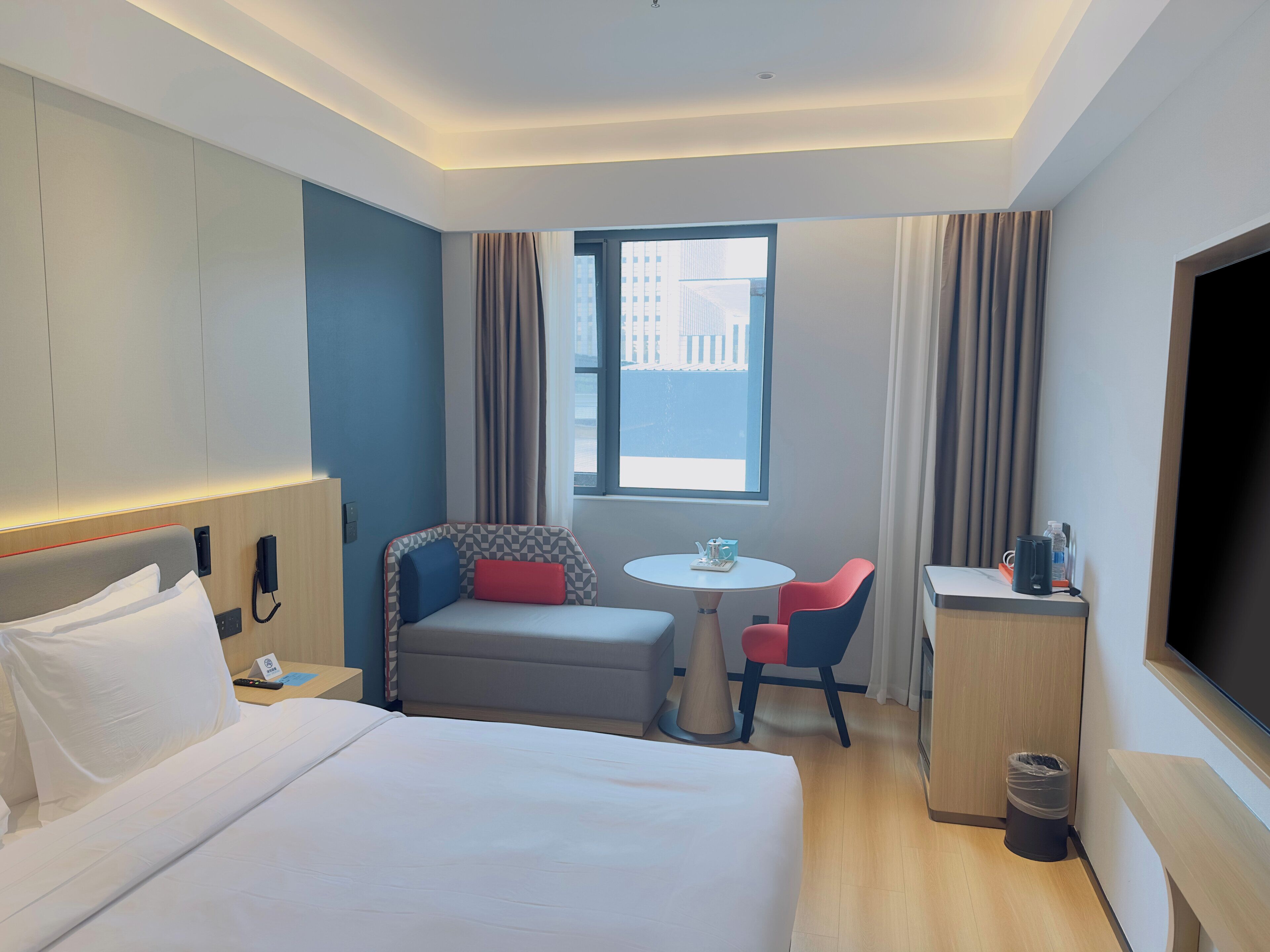 Habitación de hotel con cama, escritorio, silla, mesita, televisor y ventana con cortinas.