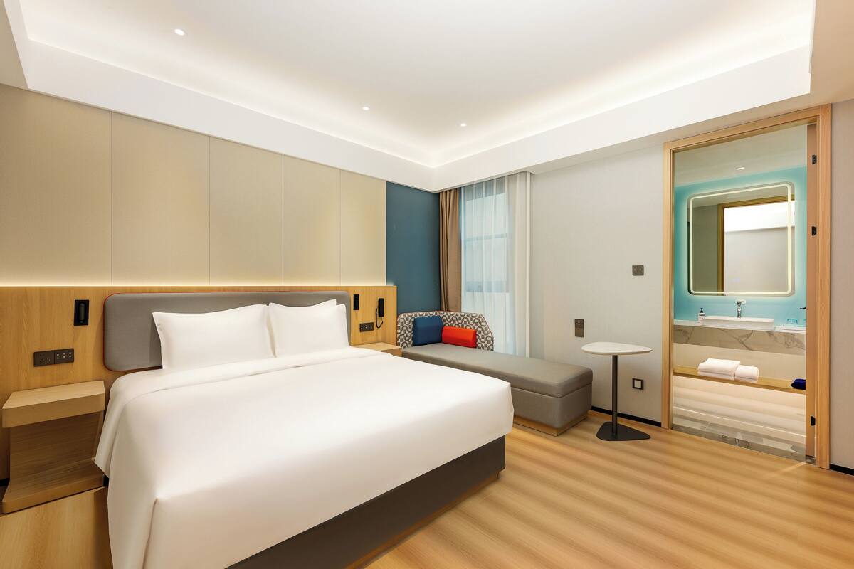 A modern hotel room with a large bed, a small sofa, a bedside table, a round table, and a bathroom with a mirror and sink.