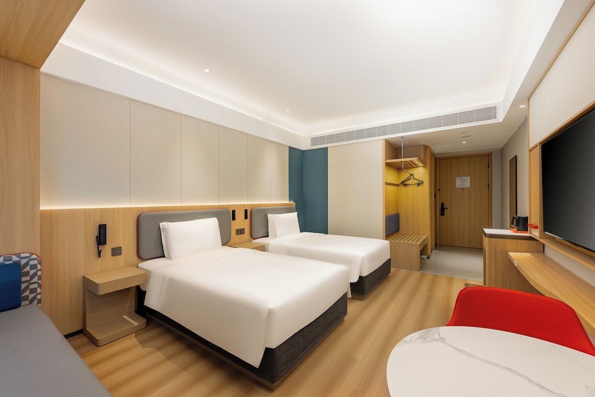 A modern hotel room with two beds, a desk, a TV, and a red chair.