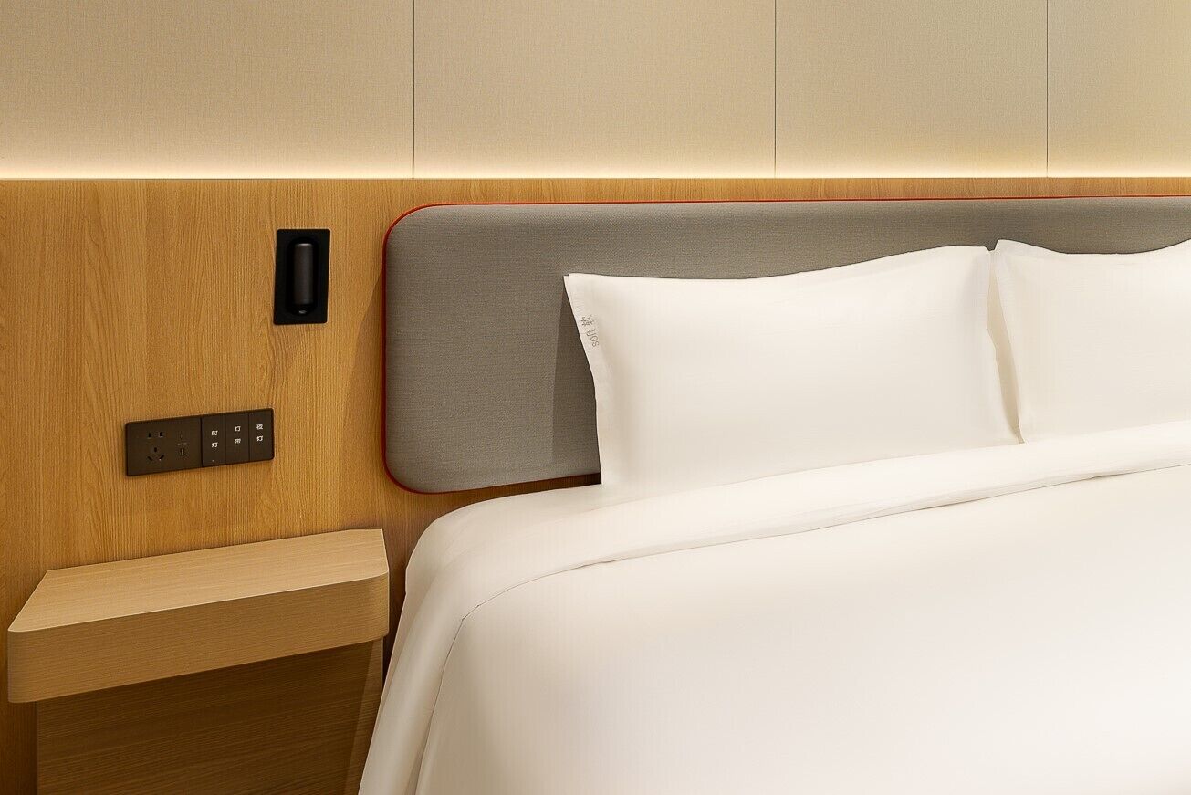 A modern hotel room with a wooden headboard, a bedside table, and a control panel.