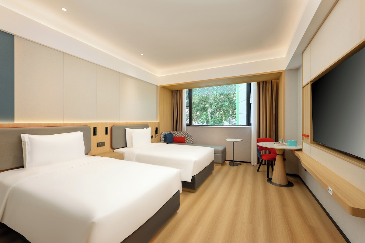A hotel room with two beds, a small table, a TV, and a window with curtains.