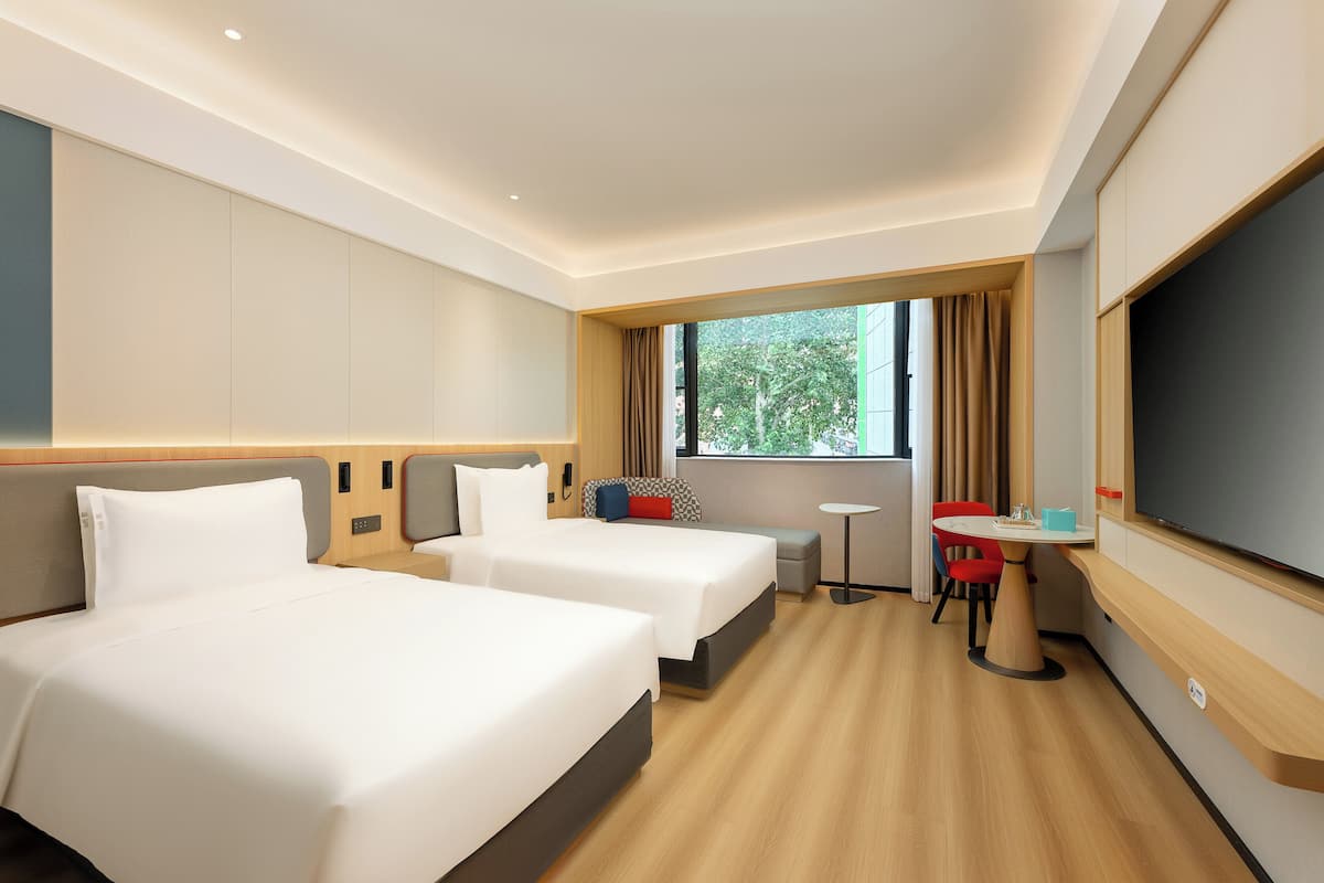 A hotel room with two beds, a small table, a TV, and a window with curtains.
