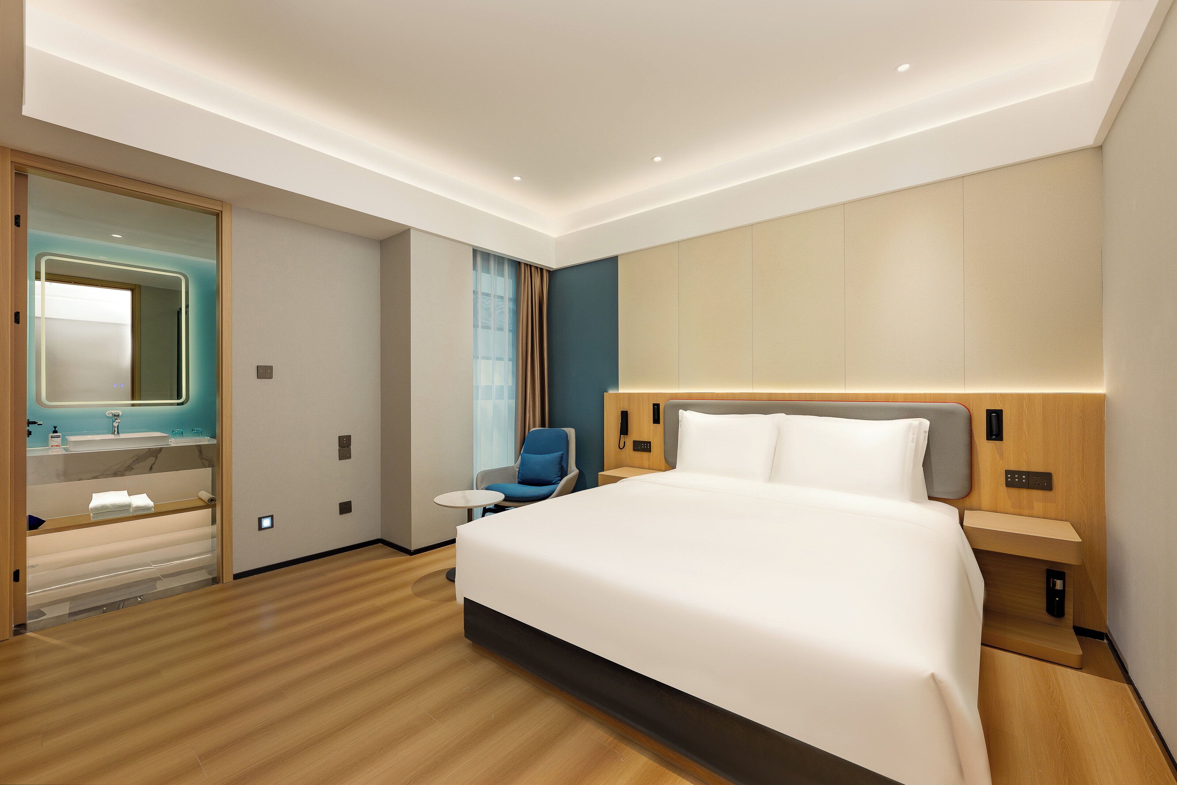 Quarto de hotel moderno com uma cama grande, mesa de cabeceira, uma cadeira, uma mesinha e um banheiro com espelho e pia.
