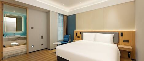 A modern hotel room with a large bed, a bedside table, a chair, a small table, and a bathroom with a mirror and sink.