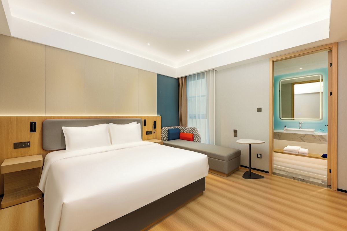 A modern hotel room with a large bed, a small sofa, a bedside table, a round table, and a bathroom with a mirror and sink.