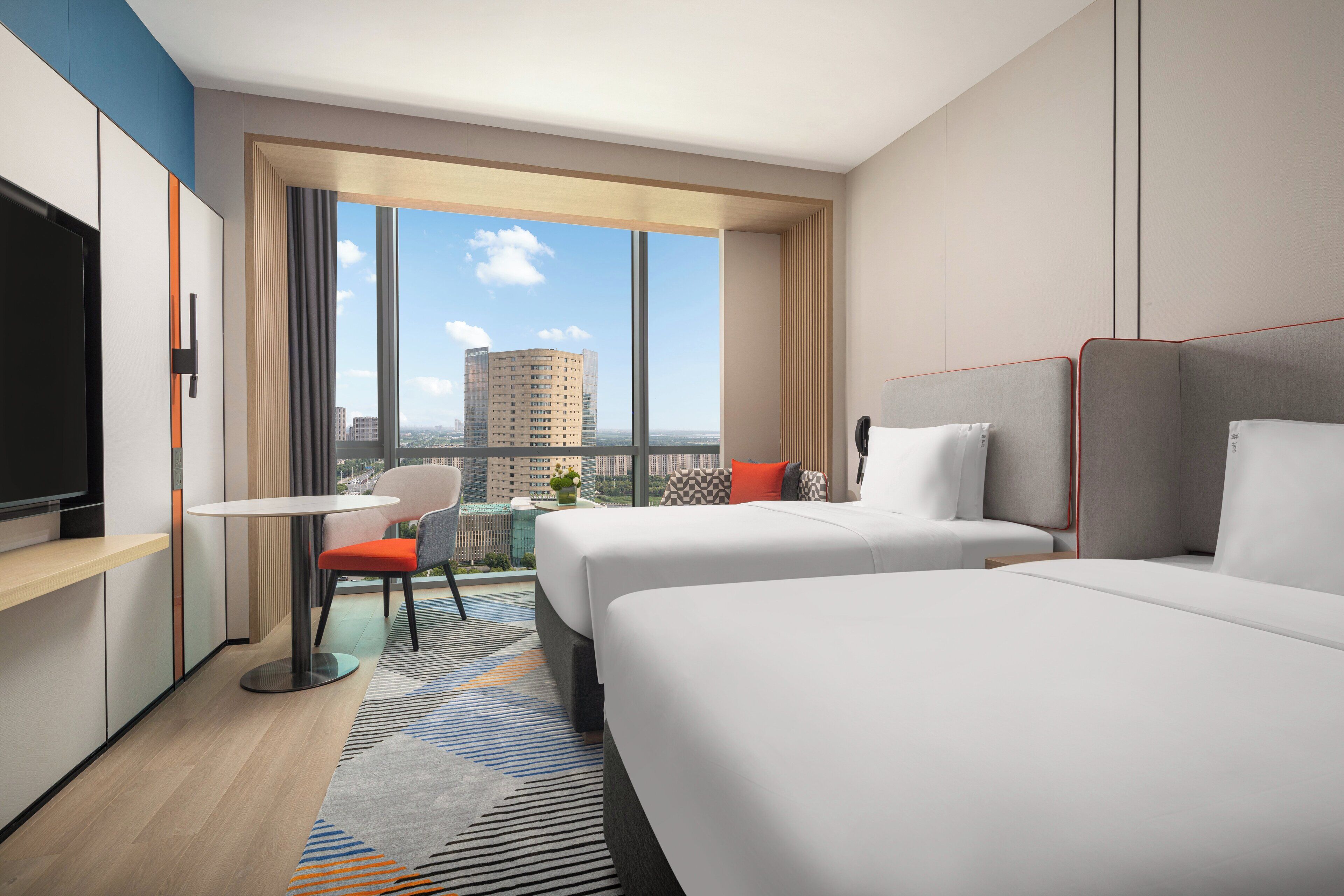 A modern hotel room with two beds, a TV, a small table, and a view of the city.