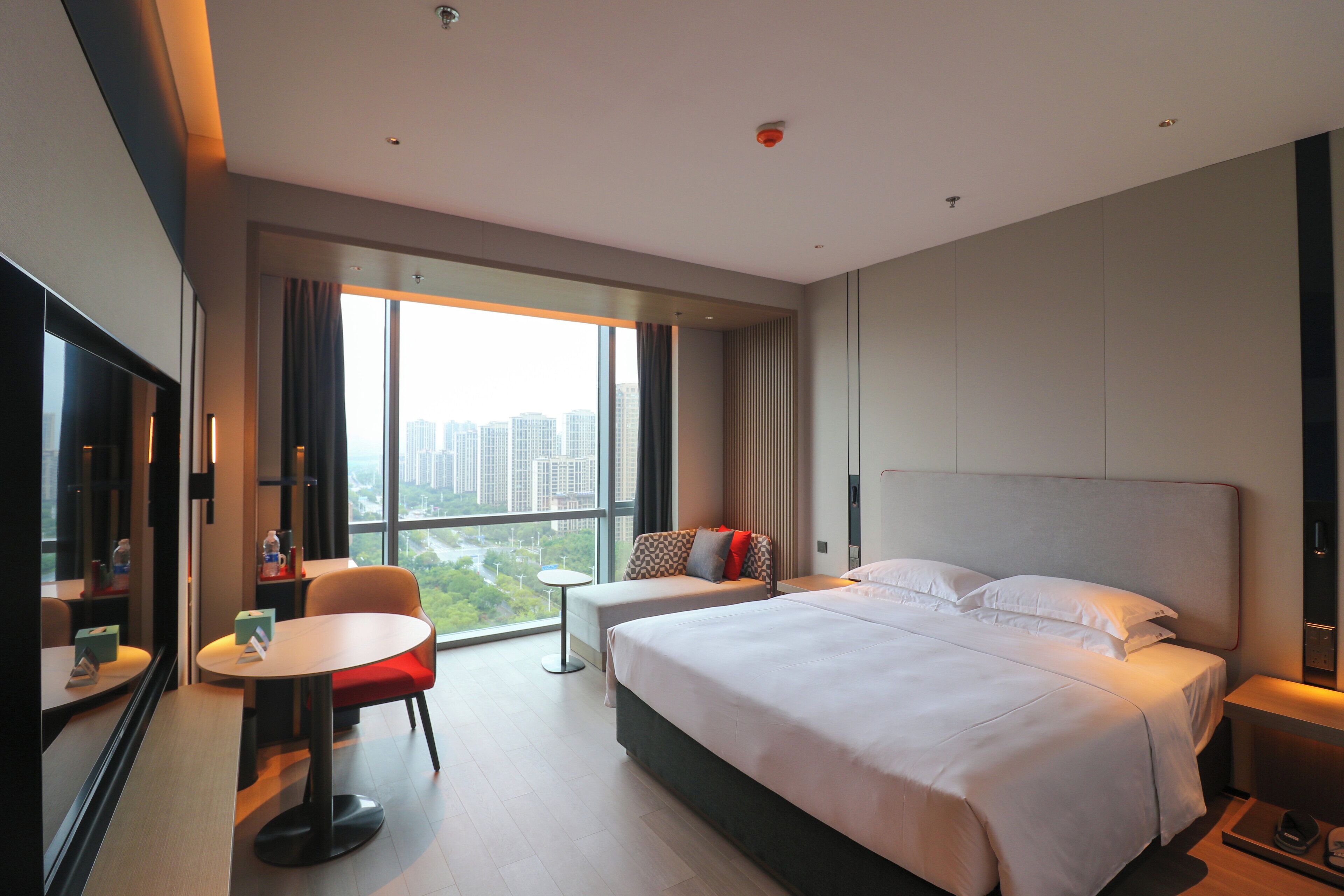 A modern hotel room with a large bed, a desk, and a chair. There is a window with a view of the city.