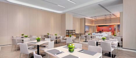 A modern hotel dining area with multiple tables and chairs, a kitchen in the background, and a clean, minimalist design.