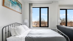 Una habitación de hotel con cama, ventana con vista a la ciudad, un cuadro en la pared y una lámpara.