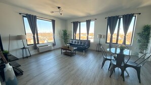 Amplia sala de estar con sofá de cuero negro, una mesita de centro y un comedor con sillas y mesa. Amplias ventanas con cortinas ofrecen vistas al exterior.