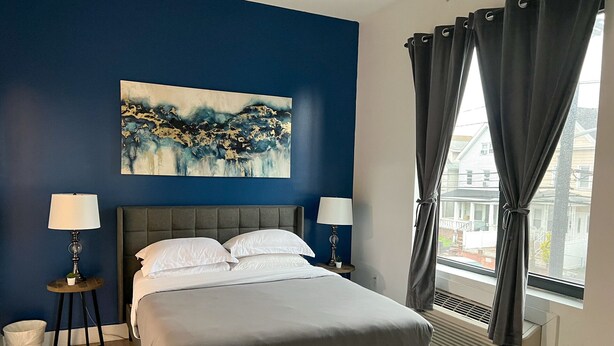 A bedroom with a bed, two bedside tables with lamps, a wall painting, and a window with curtains. - 2BR apt near EWR airport 7xw8 (Elizabeth)