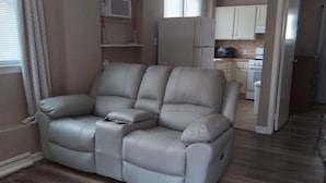 Ein Wohnzimmer mit grauem Ecksofa, einem Deckenventilator und einer Küchenzeile mit weißen Schränken und Geräten.