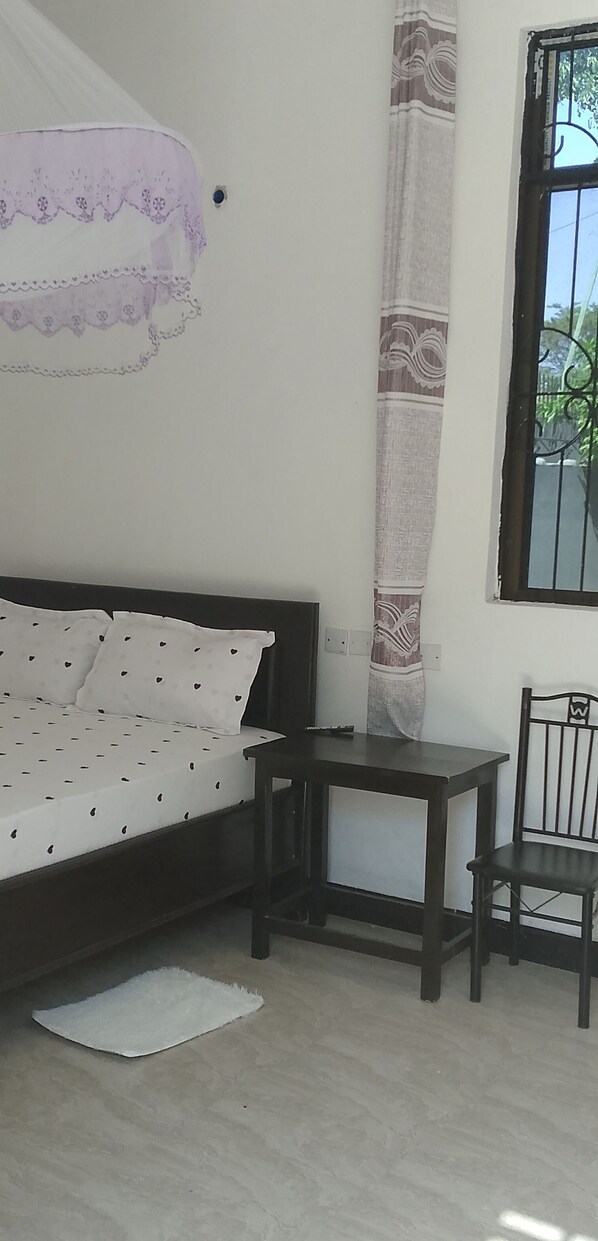 A bedroom with a bed, a small table, a chair, a window with bars, and a curtain. - Elegance Lodge &Hotel (Serengeti)