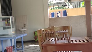 A room with a tiled floor, a wooden dining table with chairs, a blue bench, a red basket, and a window with a view of a garden.