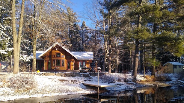 Una cabaña con un muelle que se adentra en un río tranquilo, rodeada de árboles cubiertos de nieve y un cielo despejado.