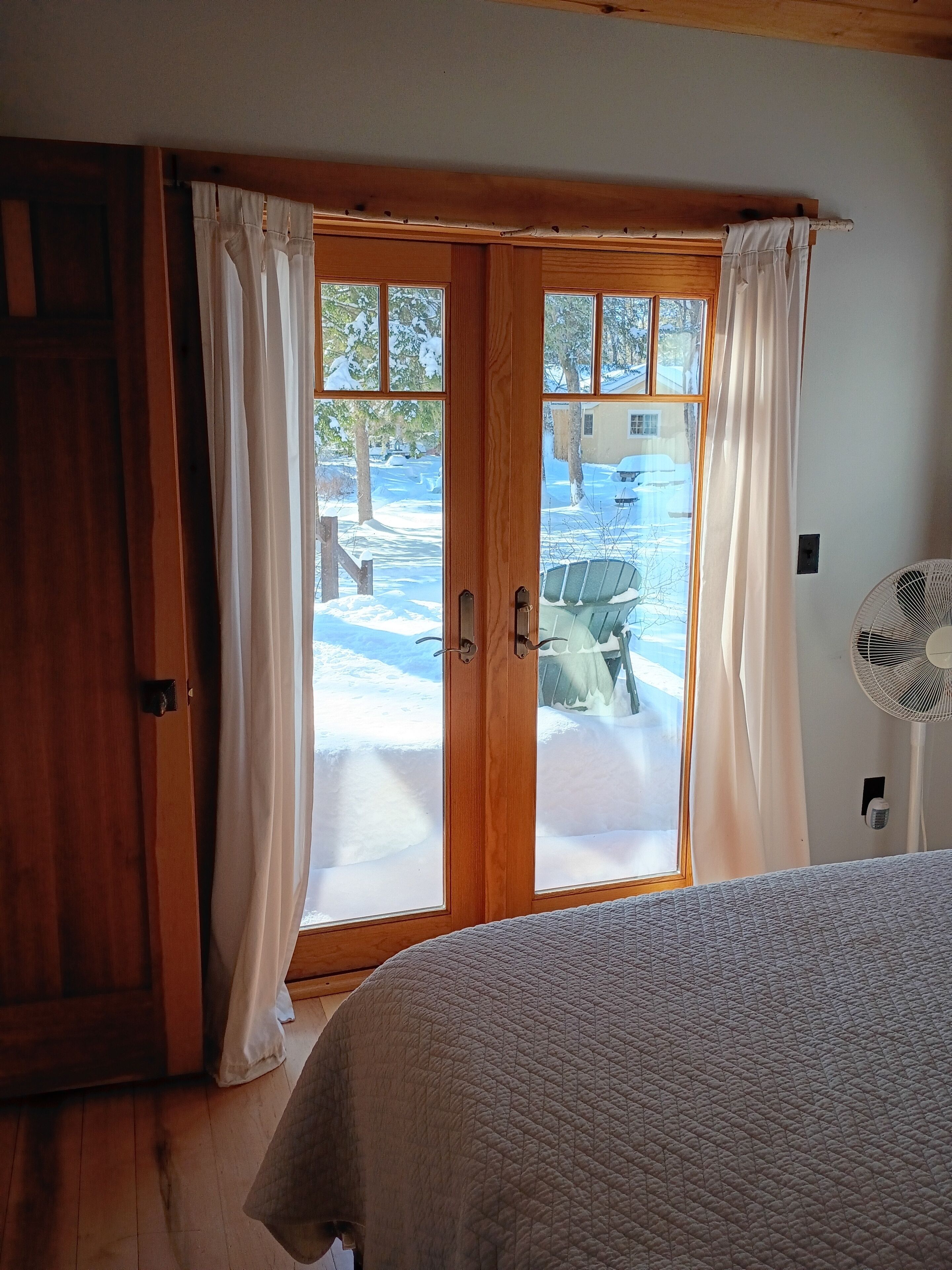 A bedroom with a wooden door, white curtains, a bed with a quilted cover, and a standing fan.