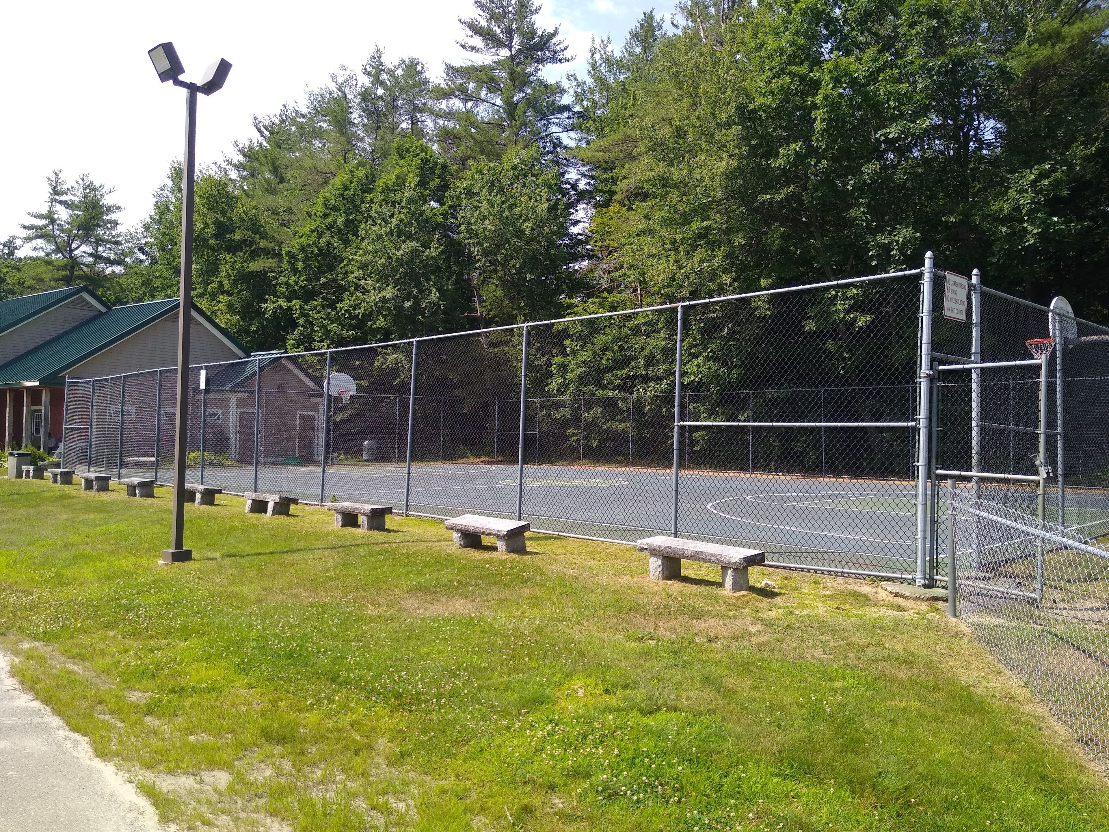 An outdoor basketball court with a chain-link fence, benches, and a building in the background.