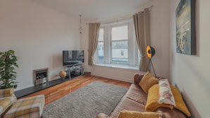 A living room with a sofa, a television, a floor lamp, and a window with curtains. - Perfect location for beach and golfing (Troon)