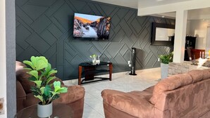 A modern living room with a large flat-screen TV mounted on a textured wall, a brown leather sofa, a glass coffee table, and a small side table with a plant. - CASA LIBERTAD 854 (Tijuana)