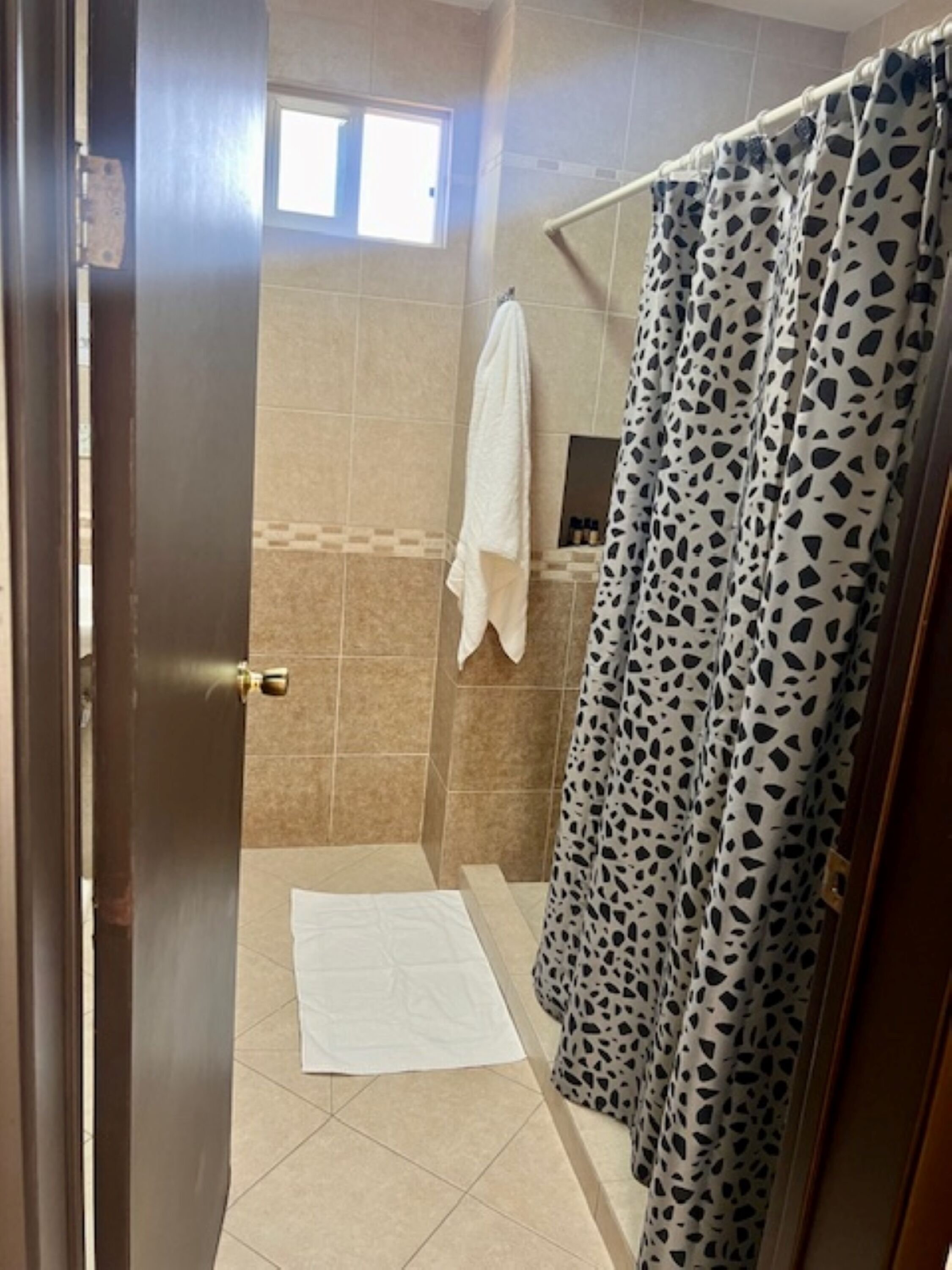 A shower area with a patterned shower curtain, a white towel hanging on the wall, and a tiled floor.