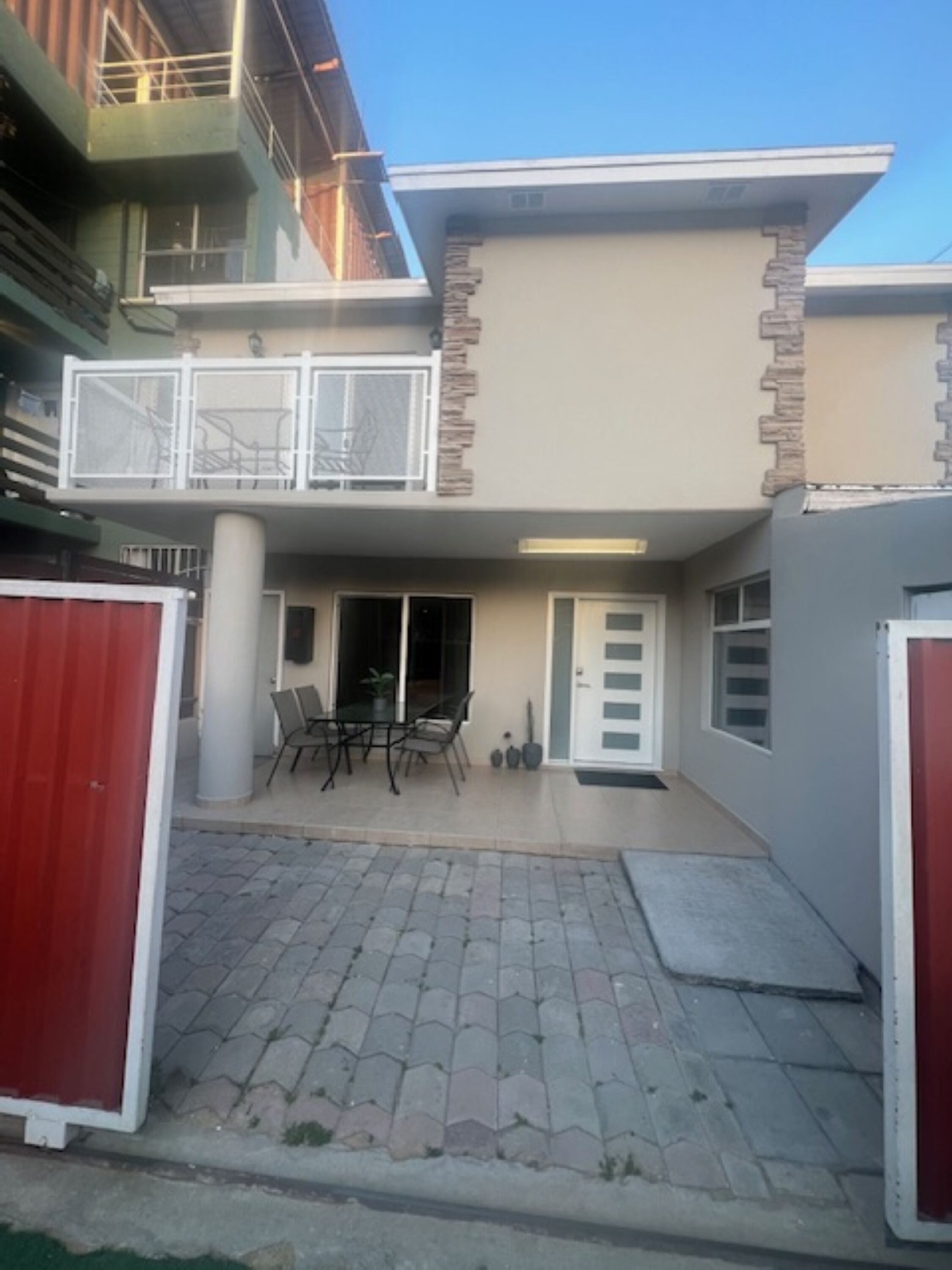 A two-story house with a balcony, a patio with a table and chairs, and a red gate.